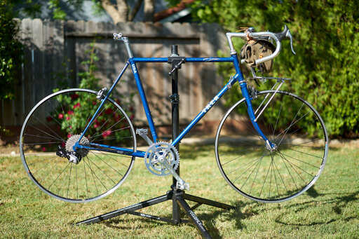 Blue bicycle on repair stand, without tires or saddle.