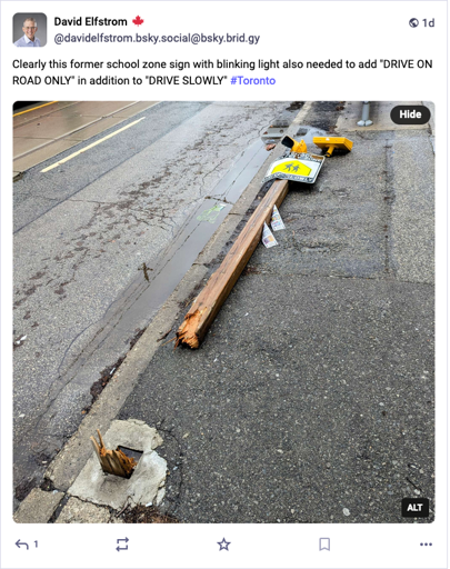 Fallen sign on a sidewalk next to a road showing the broken post. It had a blinking yellow light on top, a bright yellow triangle with walking schoolchildren silhouette and the words DRIVE SLOWLY